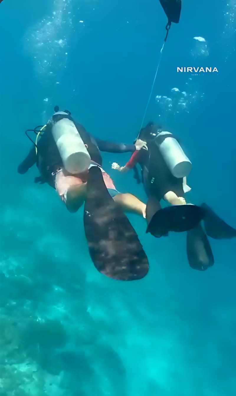 Dos buzos con tanque y aletas nadan sobre el arrecife en agua turquesa.