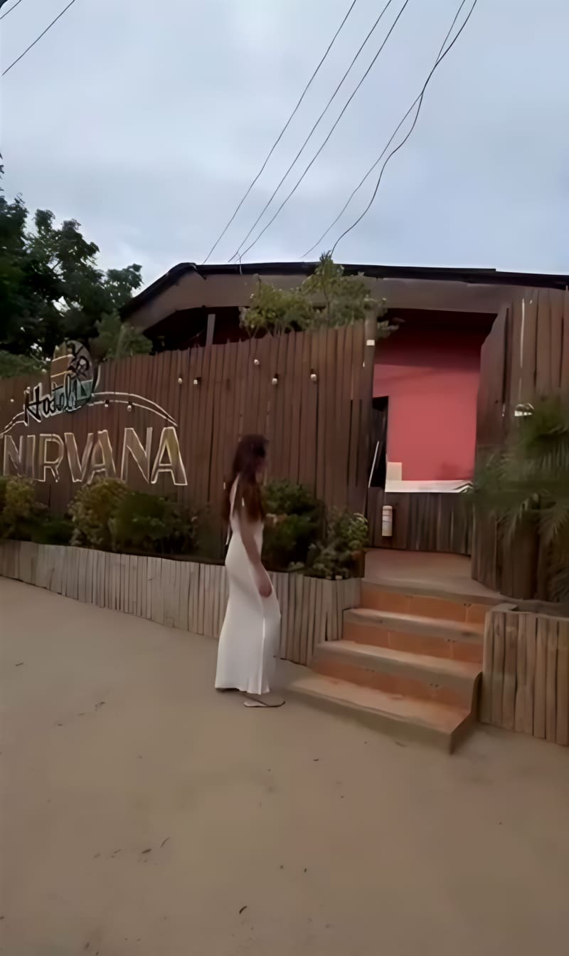 Una mujer con vestido blanco camina hacia la entrada con el letrero de madera “Hostel Nirvana”.
