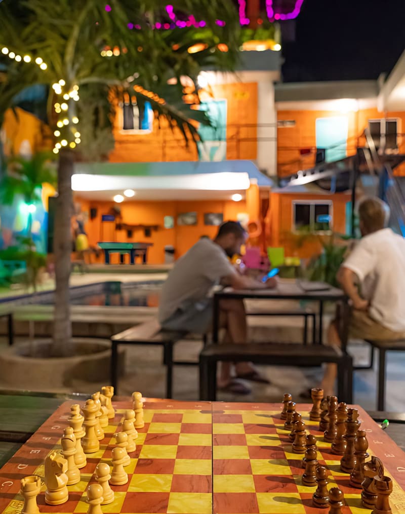 Un tablero de ajedrez en primer plano y huéspedes conversando en el patio iluminado de noche.