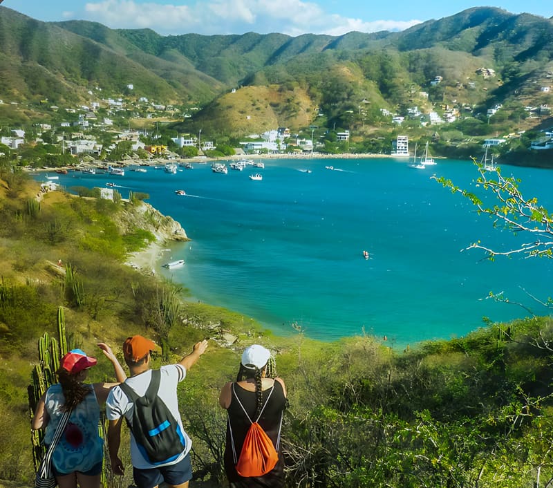 Tres caminantes miran la bahía turquesa de Taganga desde un sendero entre cactus y cerros verdes.
