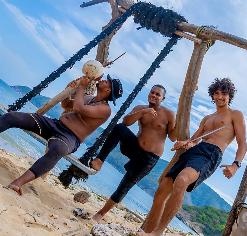 Tres chicos sonríen en un columpio de madera y cuerda en una playa, con el mar y los cerros detrás.